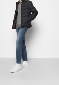 Black puffer jacket with a smooth texture, gray patterned sweater, blue jeans, and white sneakers; standing against a neutral background.