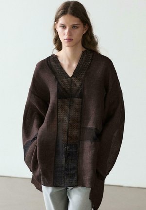 Jonge vrouw die een oversized donkerbruine gestructureerde jas met brede mouwen en lichtgrijze broek draagt, staand tegen een witte achtergrond.