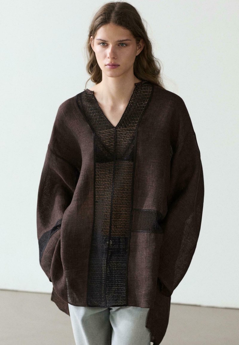 Jonge vrouw die een oversized donkerbruine gestructureerde jas met brede mouwen en lichtgrijze broek draagt, staand tegen een witte achtergrond.