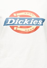 Λογότυπο Dickies με λευκό κείμενο σε μπλε πανό πάνω από κόκκινο και μπεζ σχήμα πετάλου σε λευκό ύφασμα.