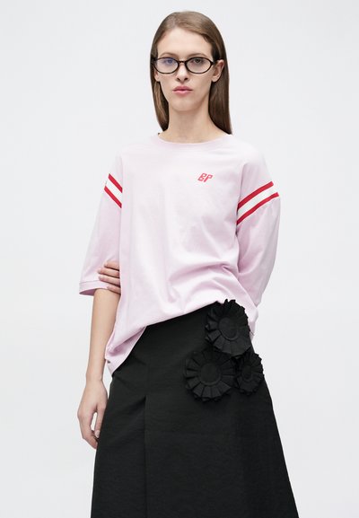 Γυναίκα με μακριά ίσια μαλλιά και γυαλιά, που φοράει ένα ανοικτό ροζ πουκάμισο σε υπερμεγέθη γραμμή με κόκκινες ρίγες και μια μαύρη φούστα με τρεις ανθισμένες διακοσμήσεις.