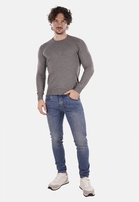 Maglione grigio a maniche lunghe, design aderente, indossato con jeans blu e sneaker chiare. Texture liscia, scollo rotondo semplice, dettagli minimi.