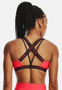Soutien-gorge de sport rose avec des bretelles élastiques noires comportant le texte "ARMOUR" et "UNDER". Les bretelles se croisent dans le dos, offrant un maintien et un soutien sécurisés.