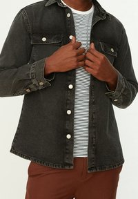 Camicia nera in denim con bottoni e due tasche sul petto con dettagli in argento, indossata sopra una t-shirt a righe bianche e blu navy. Pantaloni marroni sotto.