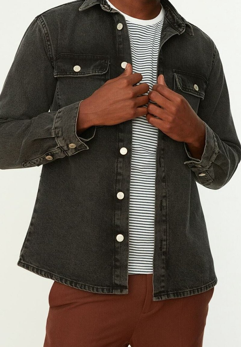 Camicia nera in denim con bottoni e due tasche sul petto con dettagli in argento, indossata sopra una t-shirt a righe bianche e blu navy. Pantaloni marroni sotto.