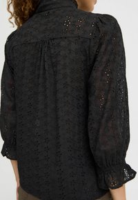 Blusa nera con un motivo floreale testurizzato, maniche a palloncino e schiena arricciata. Presenta un colletto alto e polsini elastici.