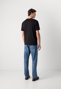 Schwarzes Baumwoll-T-Shirt mit kurzen Ärmeln, lockere Passform, schlichtes Design. Getragen mit hellblauen Denim-Jeans und schwarzen Schuhen, von hinten gesehen.