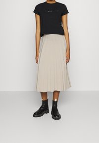 T-shirt nero corto con "DKNY" in paillettes, abbinato a una gonna midi plissettata beige e stivali alla caviglia in pelle nera con dettagli bianchi.