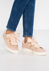 Baskets rose clair avec une finition en suède lisse, ornées de grands nœuds en tissu et de ferrures dorées, associées à un jean en denim bleu.