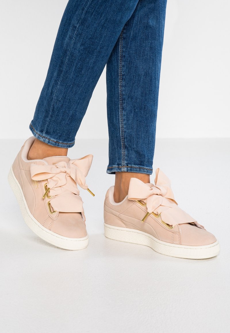 Baskets rose clair avec une finition en suède lisse, ornées de grands nœuds en tissu et de ferrures dorées, associées à un jean en denim bleu.