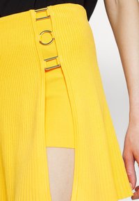 Jupe jaune côtelée avec une fente latérale, dotée d'accents métalliques circulaires argentés. Le matériau est extensible, révélant une doublure intérieure ajustée.