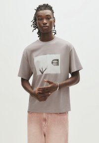 Grå bomulds T-shirt med korte ærmer, der har et grafisk motiv af et træ og en cirkulær form på forsiden, parret med lyserøde bukser.