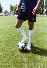 Puma nogometne tenisice s ružičastim detaljima, bijele čarape i tamnoplave kratke hlače. Igrač je spreman pored raznobojne nogometne lopte na travi.