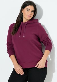 Felpa bordeaux realizzata in morbido tessuto, con cappuccio regolabile, polsini e orlo a costine, abbinata a pantaloni neri. Il modello si trova di fronte a uno sfondo bianco.