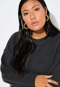 Suéter gris oscuro con puños y cuello acanalados. La modelo lleva pendientes grandes de aro dorados, con el cabello largo y oscuro, y un anillo plateado en una mano.