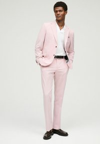 Traje rosa claro con un acabado texturizado, combinado con una camiseta polo blanca. Un cinturón negro y mocasines de cuero completan el look.