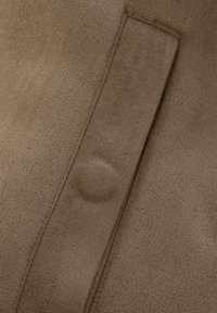 Brunt mockatygl med en mjuk, strukturerad yta. Har en synlig söm och en ficköppning, utan ytterligare beslag eller utsmyckningar.