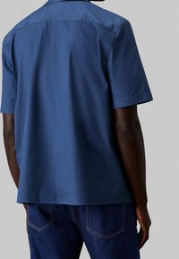 Mies, joka on pukeutunut lyhythihaiseen siniseen paitaan ja tummansinisiin farkkuihin, kuvattuna takaviistosta yksinkertaista harmaata taustaa vasten.