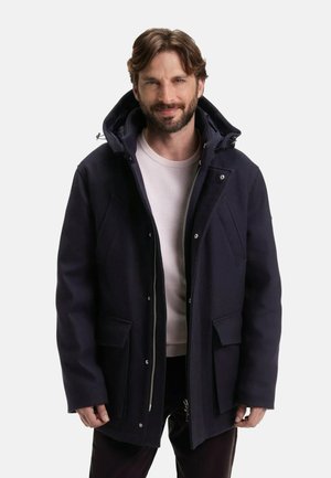 Parka en mélange de laine bleu marine avec une grande capuche, fermeture éclair et pression, deux poches avant et tissu texturé. Le modèle porte un pull léger en dessous.