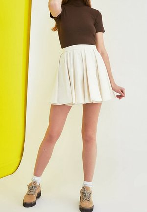Vrouw draagt een bruin kort mouw topje, witte geplooide rok, witte sokken en beige veterplateauschoenen tegen een geel-witte achtergrond.