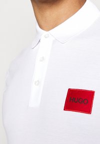 Polo blanc en maille texturée, doté d'une patte de boutonnage à trois boutons et d'un patch carré rouge avec le mot "HUGO" brodé en noir.