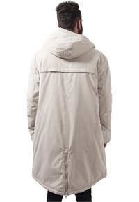 Manteau capuche beige clair avec un ourlet long et arrondi, détails de couture visibles et une fermeture éclair à l'arrière. Le tissu semble lisse et légèrement brillant.