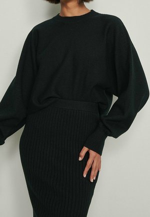 Woman wearing a black ribbed knit skirt and matching loose long-sleeve sweater, posing against a plain light background.