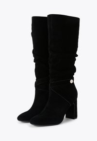 Bottes noires en daim jusqu'au genou avec un bout pointu et un talon block. Elles présentent un design froncé et une sangle décorative avec un accent circulaire.