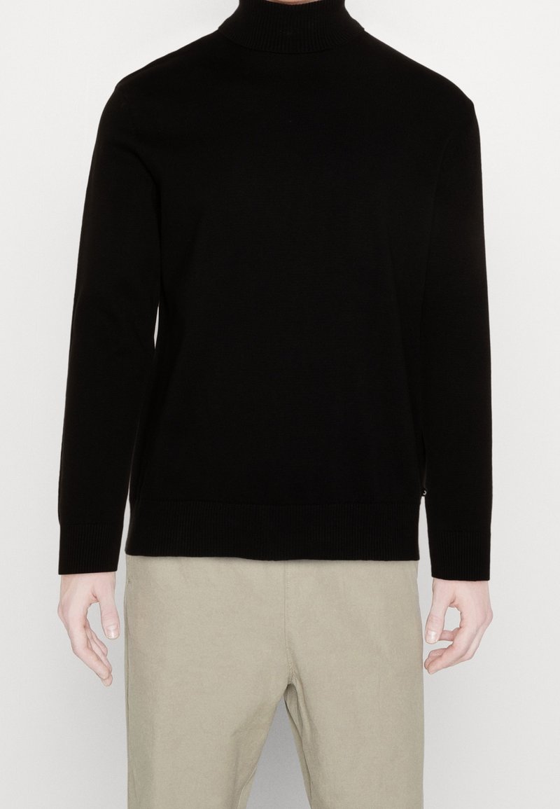 Personne portant un pull à col roulé noir et un pantalon beige, debout devant un fond clair uni, les mains relâchées le long du corps.
