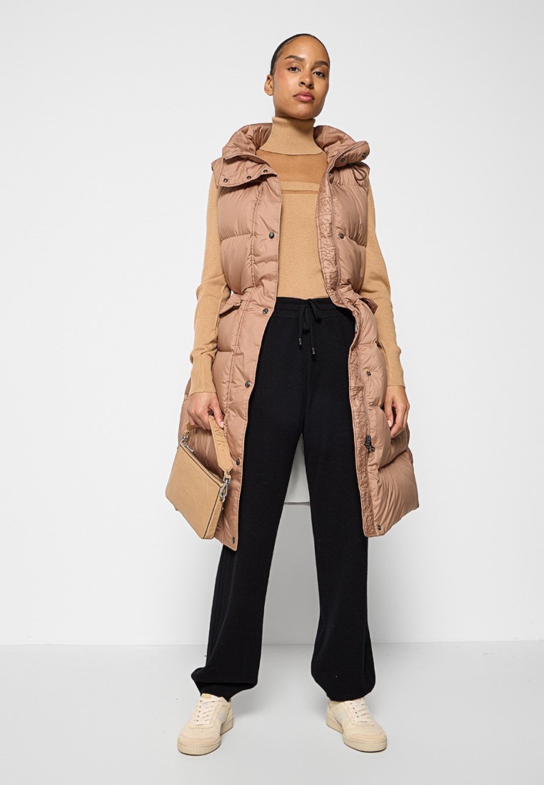 Langer, gepolsterter beiger Weste mit Kapuze, Frontknöpfen und Seitentaschen, getragen über einem engen beigen Oberteil und schwarzen Jogginghosen, während eine beige Handtasche gehalten wird.