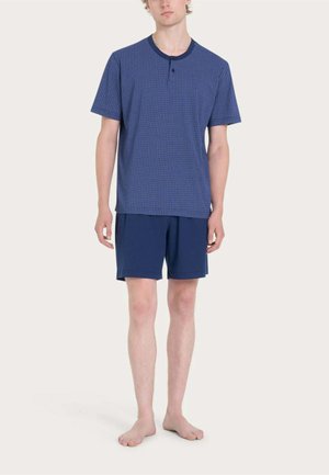 Uomo in piedi a piedi nudi che indossa una maglietta henley blu a maniche corte con motivo e pantaloncini blu semplici su uno sfondo semplice.