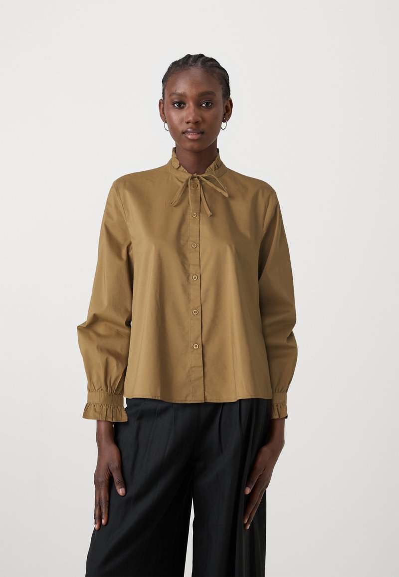 Chemise à manches longues beige clair avec un col froncé, un devant boutonné et un détail de nœud, associée à un pantalon sombre. Tissu lisse et ajusté.
