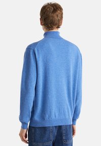 Muž v světle modrém roláku a modrých džínách, stojící zády k fotoaparátu na jednoduchém bílém pozadí.