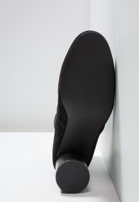 Botte noire à talon vue de la semelle avec un talon circulaire texturé reposant contre une surface d'angle blanche.
