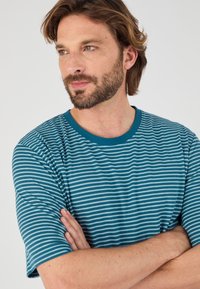 Camiseta a rayas en verde azulado y azul claro, hecha de un tejido suave con cuello redondo y mangas cortas; ajuste casual con rayas horizontales visibles.