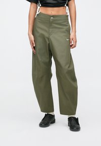 Pessoa vestindo calças largas verdes-oliva, ténis pretos e um top cropped preto com detalhes brancos em cordão, de pé contra um fundo simples.