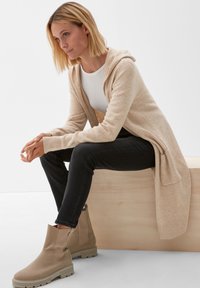 Beige kardigan s kapucí a dlouhými rukávy, s žebrovanými manžetami, spojený s černými džíny a krémovými kotníkovými botami na dřevěném povrchu.