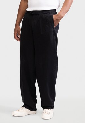 Άνδρας που φοράει φαρδιά μαύρη παντελόνα με πλαϊνές τσέπες και λευκά αθλητικά, στέκεται μπροστά σε απλό φόντο.