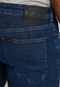 Dark blue denim jeans featuring a black leather waistband label with "Lee" embossed. Includes a back pocket with a small branded tag.
