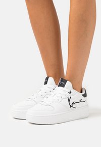 Hvide sneakers med en glat læderfinish, sorte detaljer på hælen og et markant signaturdesign på siden. Klassisk snørestil.