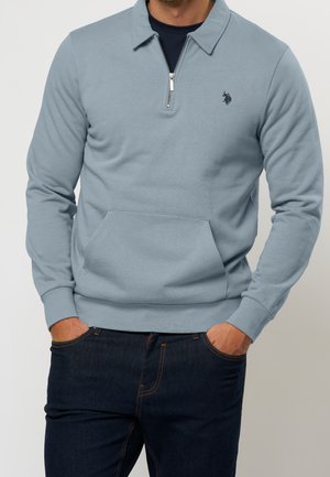 Mand iført lyseblå quarter-zip pullover med forlomme og mørkeblå jeans, hænderne i lommerne, synlig fra nakke til hofter.