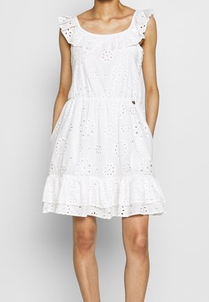 Femme portant une robe blanche sans manches en tissu ajouré, avec un col à volants et un ourlet à volants, les mains dans les poches latérales.