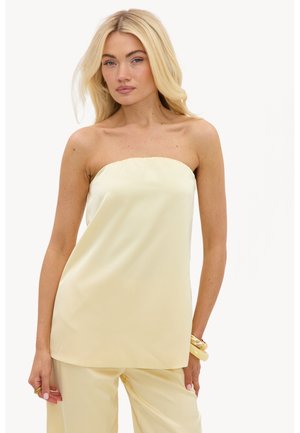 Femme aux longs cheveux blonds portant un haut jaune pâle sans bretelles et un pantalon assorti, avec des bracelets dorés au poignet droit.