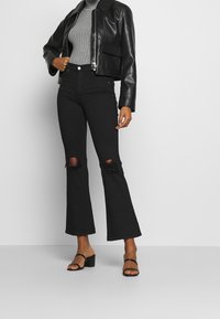 Femme portant un jean flare court noir avec genoux déchirés, des sandales noires à talons, un col roulé gris côtelé et une veste en cuir noire.