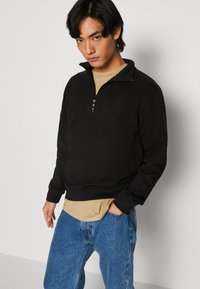 Sort quarter-zip sweatshirts med ribbede ærmer og kant, smidt sammen med en tan farvet skjorte og blå jeans. Blødt stof med en struktureret overflade.