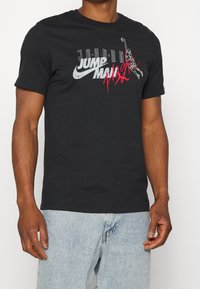 Musta puuvillapaita, jossa on valkoinen ja punainen "Jordan Jumpman" -grafiikka. Lyhyet hihat, pyöreä kaula-aukko, rento malli, sileä pinta.