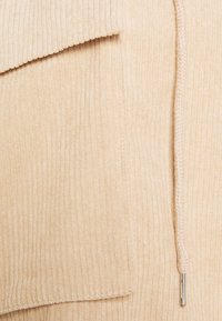 Beige gerippter Stofftextur mit einer Taschenklappe und Kordelzugdetails mit einem metallischen Endstück. Weiche und strukturierte Oberfläche.