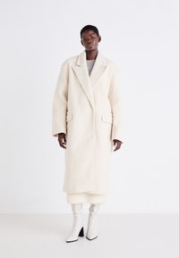 Long manteau ivoire en tissu doux et texturé avec un large col et de grandes poches avant, associé à des bottes blanches brillantes montant jusqu'aux genoux.