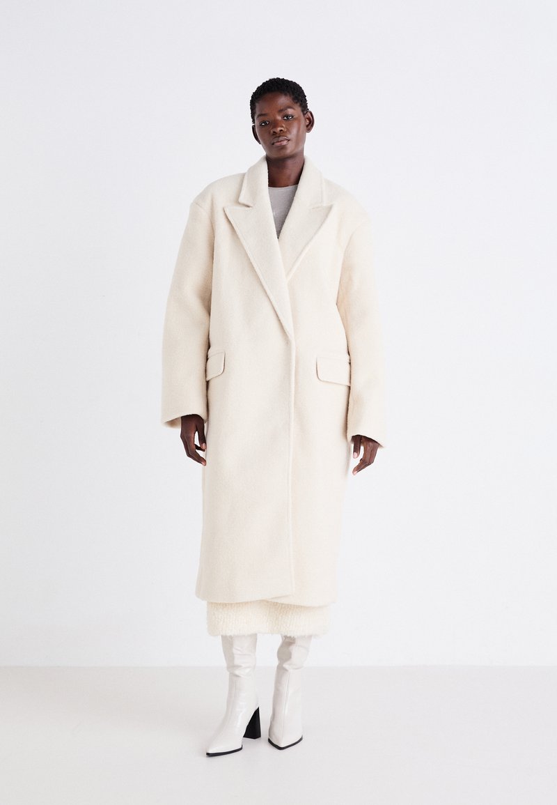 Long manteau ivoire en tissu doux et texturé avec un large col et de grandes poches avant, associé à des bottes blanches brillantes montant jusqu'aux genoux.