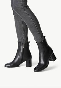 Czarne skórzane botki za kostkę na słupku, z bocznym zamkiem błyskawicznym i elastycznymi panelami. Gładka texura i elegancki design uzupełniają szare dżinsy.